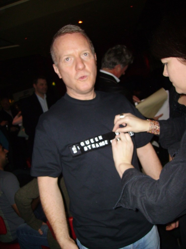 A photo of a woman pinning something onto a man's t-shirt, with writing across the front. The man is looking up towards the ceiling. 