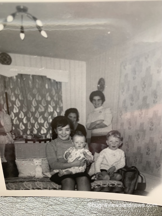 An old 1960s black and white photo of a woman sat on a sofa with a baby in her arms and a small child sat next to her. Behind the sofa is standing woman and an older woman who is sat down.