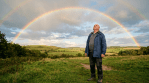 Man standing in a grassy field looking up at a double rainbow in a partly cloudy sky