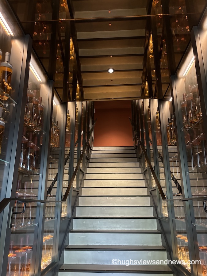 A low-angle view looking up a modern indoor staircase with light-coloured steps leading toward a warm, terracotta-orange wall at the top landing. The staircase is lined on both sides with tall, glass-fronted display cases illuminated with warm accent lights, showcasing rows of amber liquor bottles. Reflective, polished ceiling panels above mirror the staircase and surrounding lights