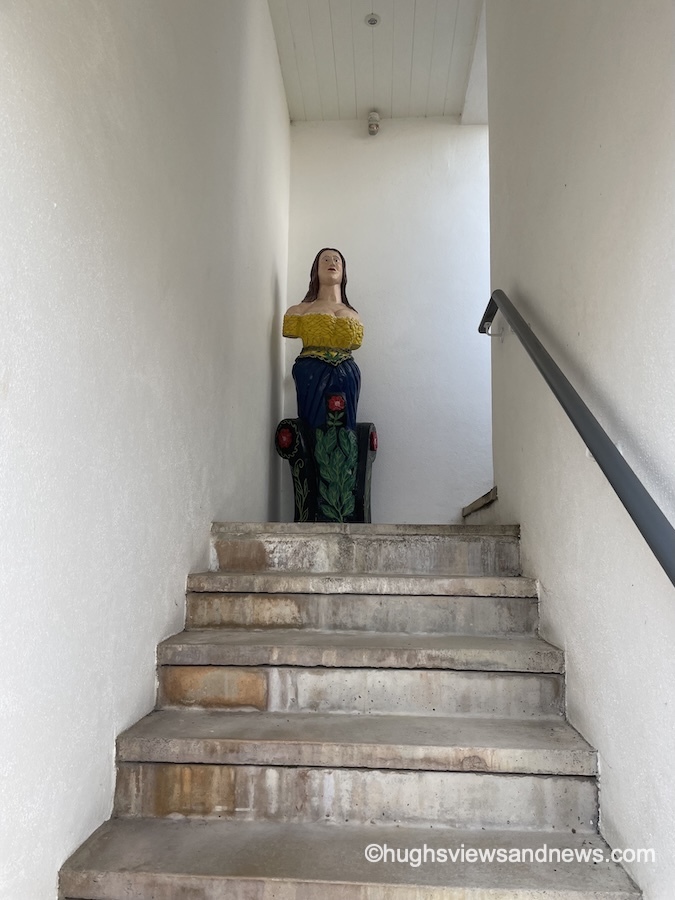 A low-angle view looking up a narrow flight of outdoor concrete steps flanked by plain white walls. At the top landing sits a colourful, carved wooden ship's figurehead of a woman wearing a yellow top and dark blue skirt. A dark metal handrail runs along the wall on the right side of the staircase.
