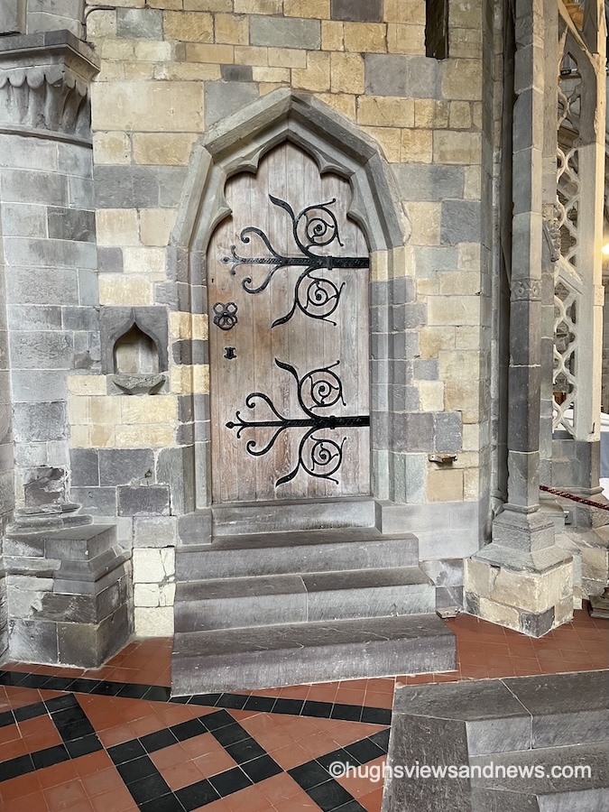 An arched, rustic wooden door set into an ancient stone wall inside a historical building or church. Four stone steps lead up to the door, which is embellished with ornate, decorative black wrought-iron hinges featuring swirling scrollwork. The floor below consists of red and black tiled patterns. In the bottom-right corner, a small text watermark reads "@hughsviewsandnews.com".